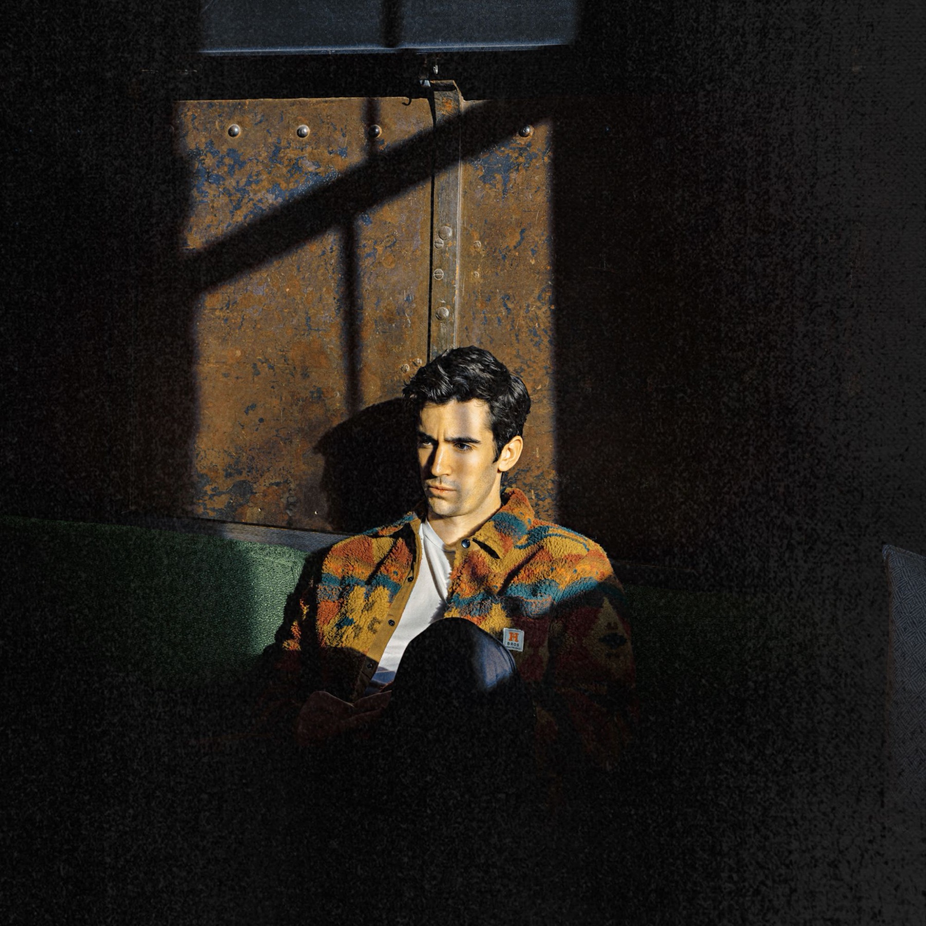 Nick Gerard sitting on a green couch in a patterned fleece jacket, lit by a shaft of sunlight against a rusted metal wall